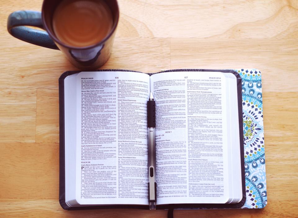An open Bible on a wooden table beside a mug of coffee and a pen
